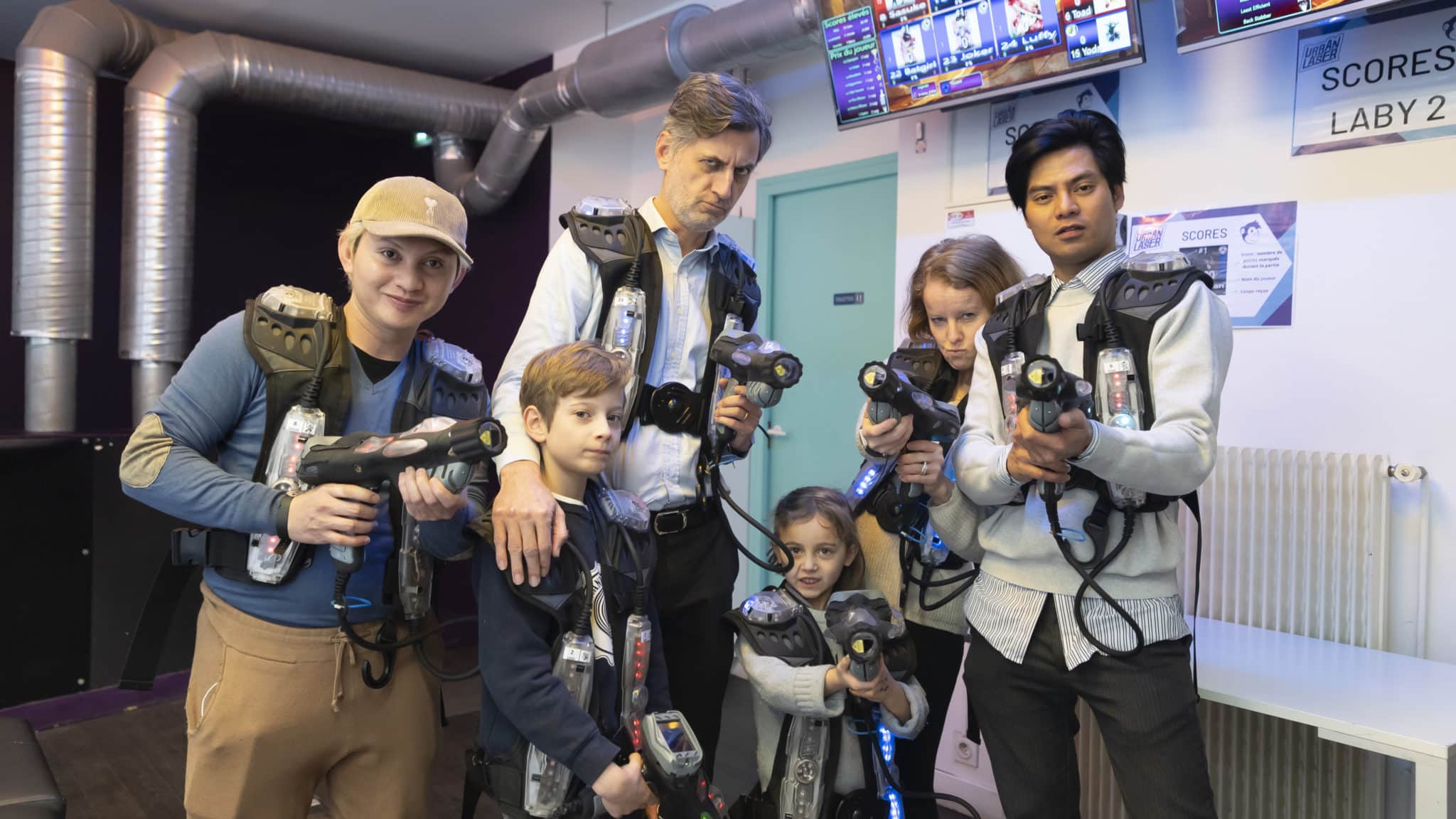 Un groupe de six personnes, dont deux enfants, équipés de gilets et pistolets de laser game, posent dans l'aire de jeu. Texte: "SCORES LABY 2".