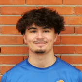 Jeune homme aux cheveux bruns foncés et au collier de barbe souriant, portant un t-shirt bleu, devant un mur de briques rouges.
